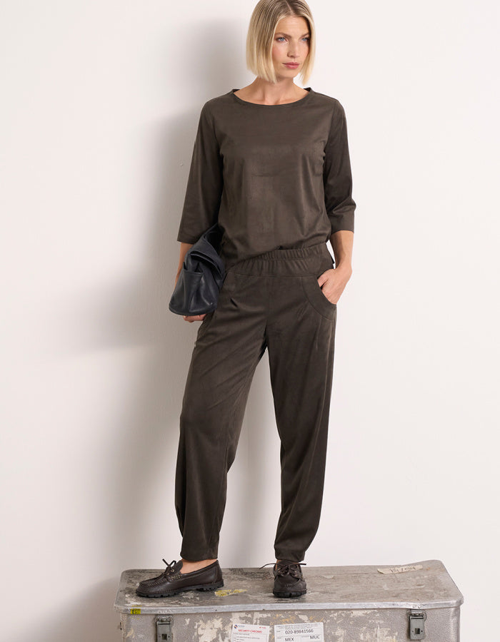 A woman models a olive two-piece lounge set consisting of a long-sleeve top and matching jogger pants, standing on a weathered metal storage box against a plain white wall.