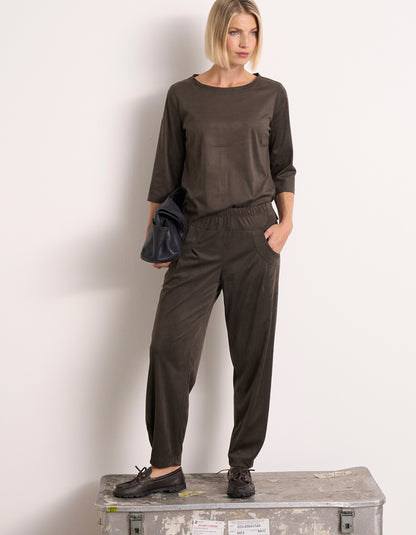 A woman models a olive two-piece lounge set consisting of a long-sleeve top and matching jogger pants, standing on a weathered metal storage box against a plain white wall.