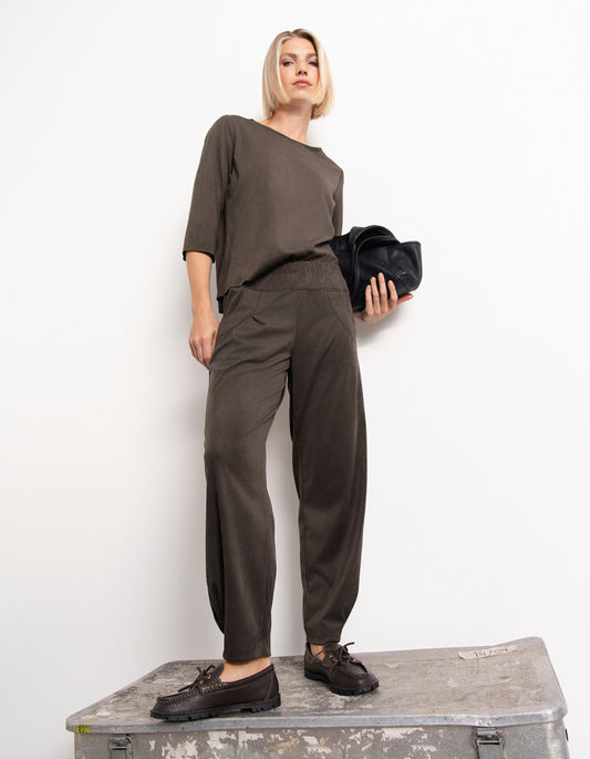 A woman modelling a olive two-piece lounge set consisting of a wide-neck top and matching relaxed trousers, standing on a weathered metal trunk against a plain white wall.