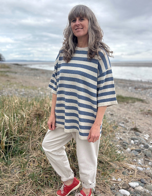 Woman wearing a Breton striped organic cotton sweater and white pants standing in a field with a beach in the background.