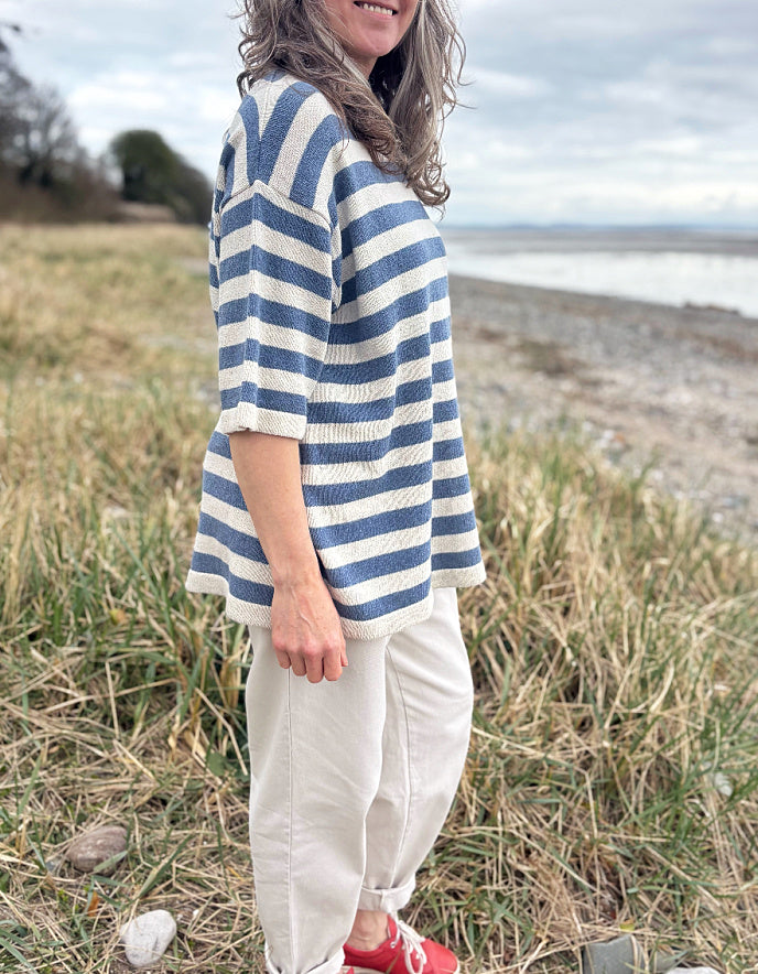 Woman wearing a Breton striped organic cotton sweater and white pants standing in a field with a beach in the background.