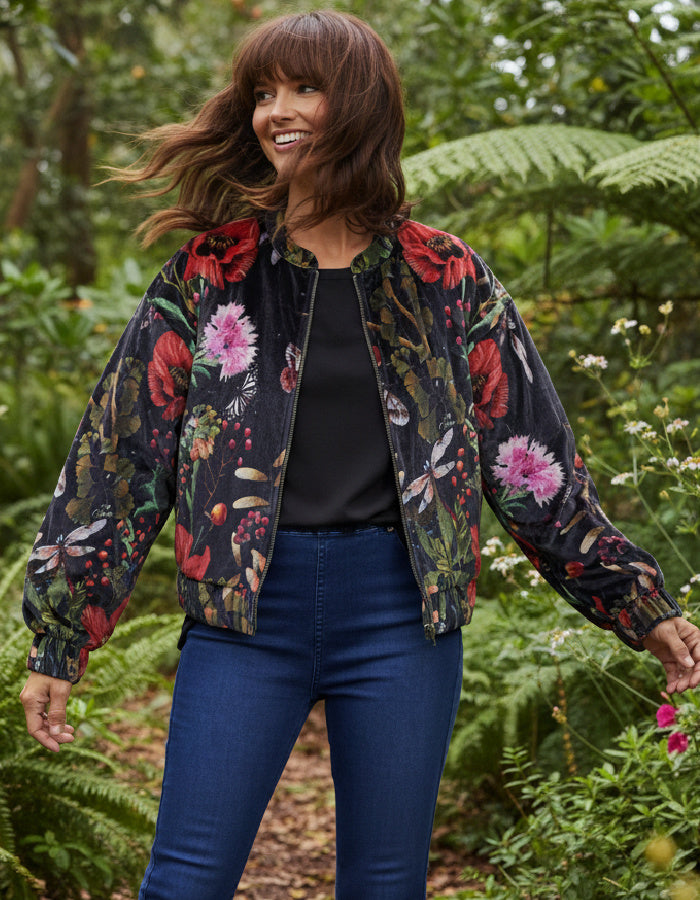 A woman wearing a black bomber jacket with a vibrant red, pink, and green floral print, styled with a black top and blue jeans, standing in a lush green garden.