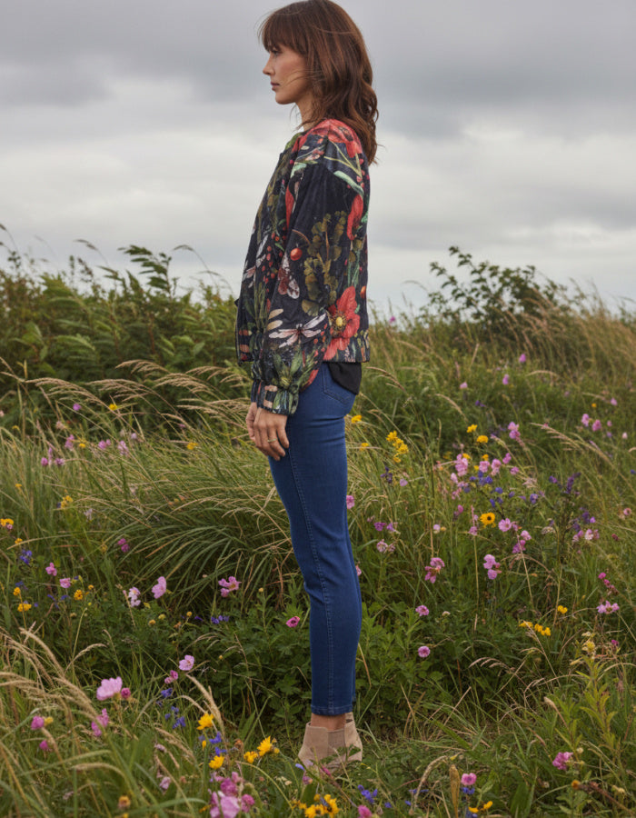 A woman stands in profile in a grassy field of wildflowers, wearing a dark floral bomber jacket, blue skinny jeans, and tan ankle boots under an overcast sky.
