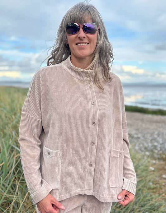 Person wearing a winter white jumbo cord jacket and sunglasses on a beach with grass and water in the background