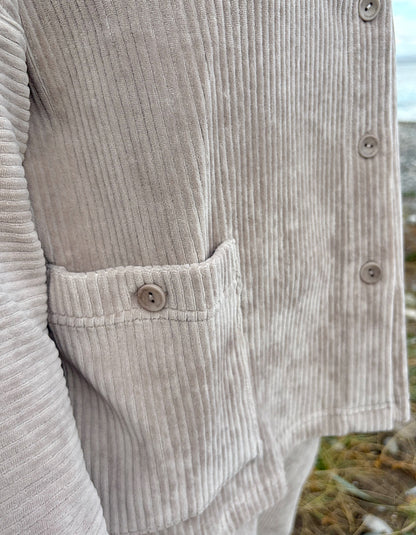 Person wearing a winter white jumbo cord jacket and sunglasses on a beach with grass and water in the background