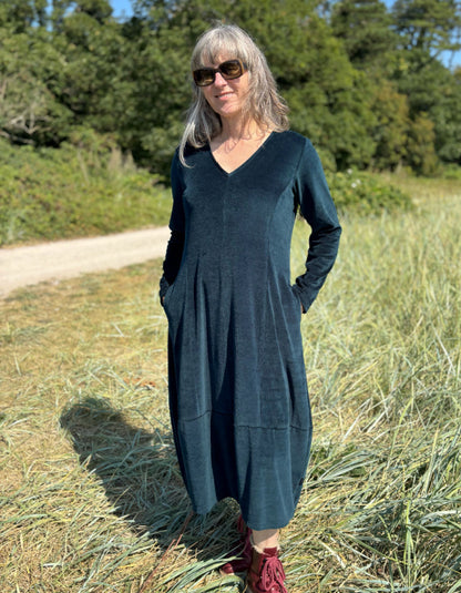 Woman in a long dark teal velvet dress with long sleeves and V neck standing in a field with trees in the background