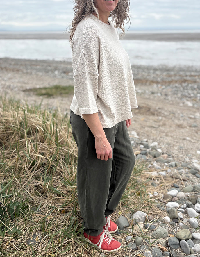 Woman standing outdoors in a natural setting wearing a cream organic cotton sweater and dark green tercel pants.