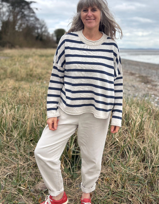 Woman wearing a Breton striped organic cotton sweater and white pants standing in a field with a beach in the background.