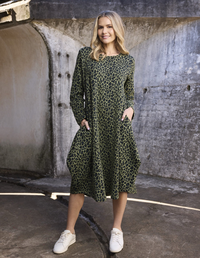 A woman modeling a long-sleeve, knee-length dress with an olive green and black leopard print, standing in front of a concrete wall.