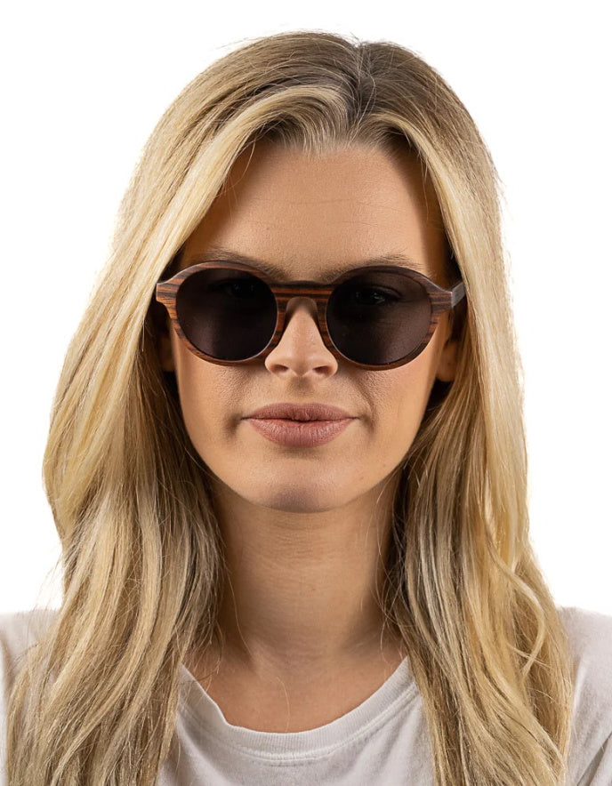A close-up of a woman with long blonde hair wearing round dark wood sunglasses with a brown acetate frame, set against a plain white background.