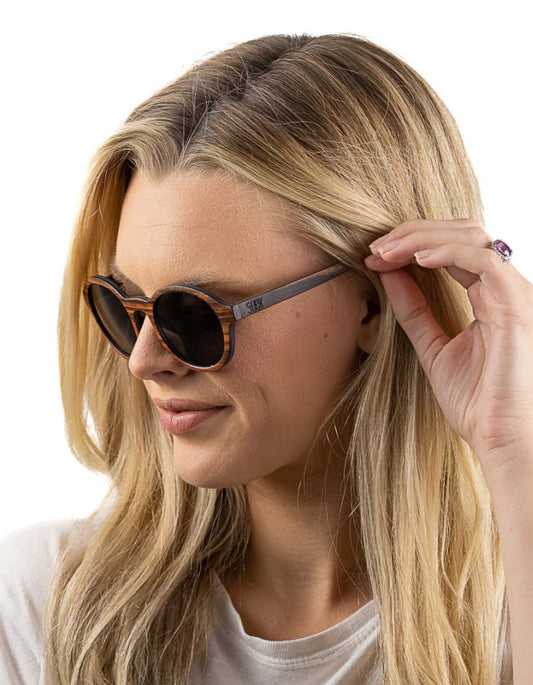A woman with blonde hair wearing round sunglasses with wood frames and dark lenses, shown in a close-up profile against a white background.