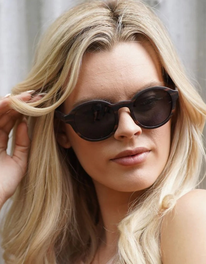 A close-up of a blonde woman wearing dark round oak wood sunglasses, with her hand touching her hair against a soft grey background.