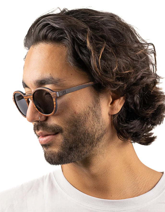 A man with dark wavy hair and a beard wearing round wood sunglasses with a brown wood-grain frame and dark lenses, shown in profile against a white background.