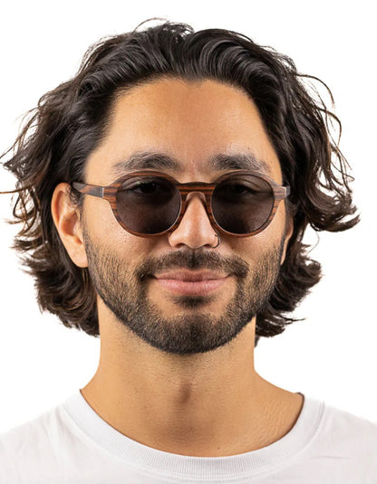 A man with wavy dark hair and a beard wearing round dark brown wood sunglasses with a translucent brown acetate frame, set against a plain white background.