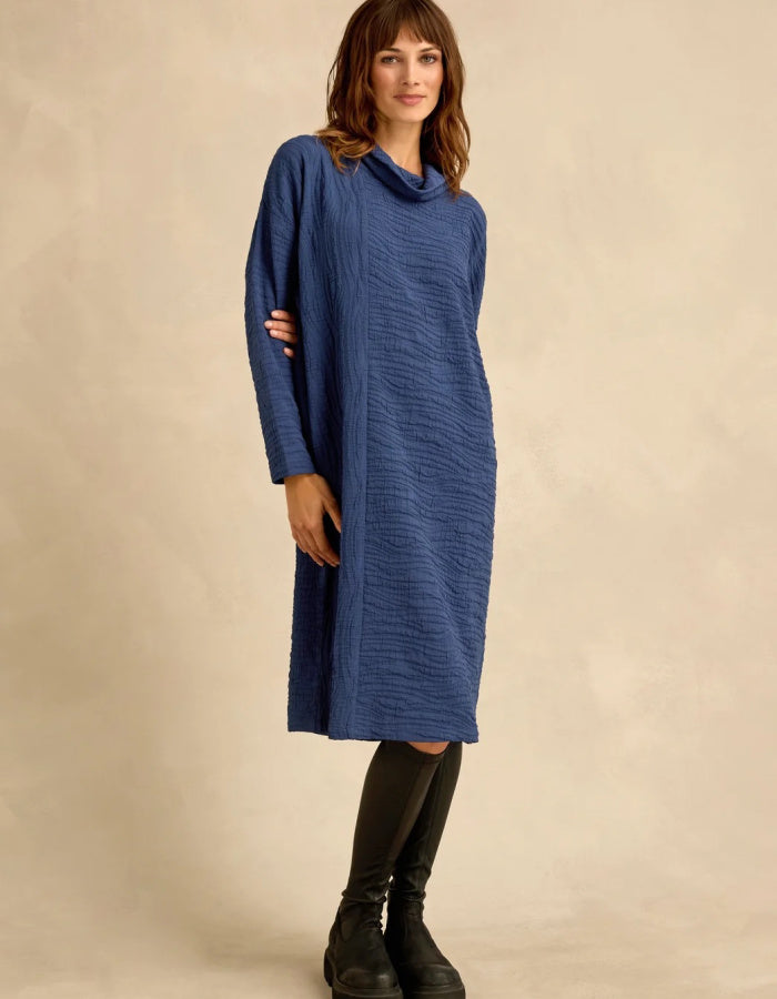A woman modeling a textured, cowl-neck midi dress in a dusty blue color, styled with black knee-high boots against a neutral beige studio backdrop.