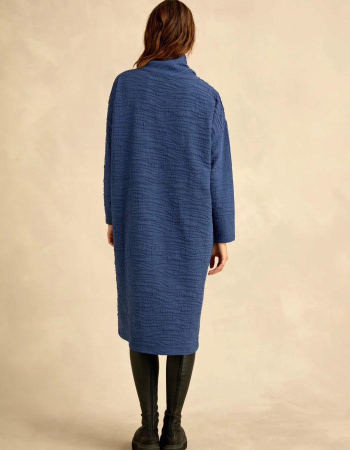 A woman viewed from behind wearing a textured, crinkled blue long-sleeve coat over dark leggings and boots, standing against a warm beige studio backdrop.