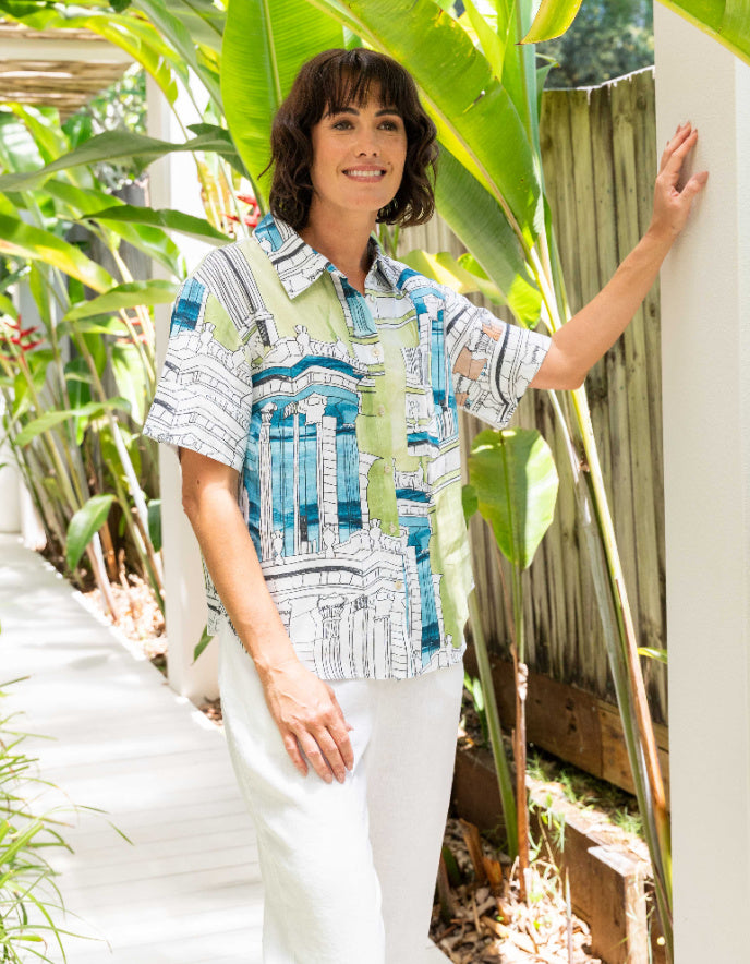 Woman wearing a patterned shirt and white pants standing outdoors with greenery around.