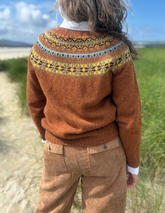 A person viewed from behind wearing a rust-orange knit sweater with a colorful Fair Isle yoke pattern in yellow, blue, and brown, paired with brown corduroy trousers, standing outdoors on a sandy path with grass and sky in the background.