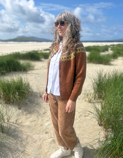 A woman standing on a sandy beach dune path wearing a brown knit cardigan with a colorful patterned yoke, a white button-up shirt, brown corduroy pants, and white sneakers.
