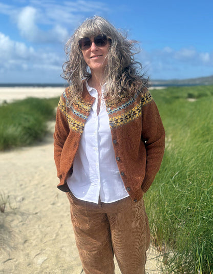 A woman with long gray hair and sunglasses stands outdoors on a sandy path near grassy dunes under a blue sky, wearing a rust-brown cardigan with a colorful geometric pattern across the chest over a white button-up shirt and brown corduroy pants.