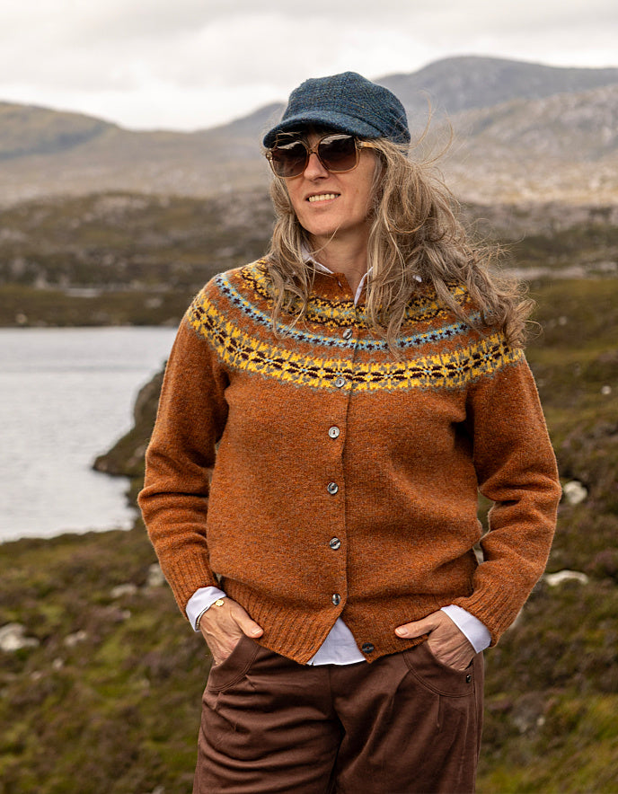 A woman wearing a brown Fair Isle knit cardigan, brown trousers, a blue cap, and sunglasses stands outdoors in a mountainous, lakeside landscape.