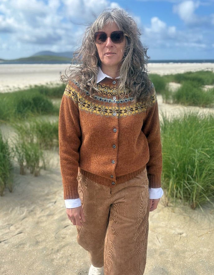 A woman with long gray hair and sunglasses stands on a sandy beach path wearing a rust-brown Fair Isle cardigan over a white collared shirt and brown corduroy trousers, with dunes and a cloudy blue sky in the background.