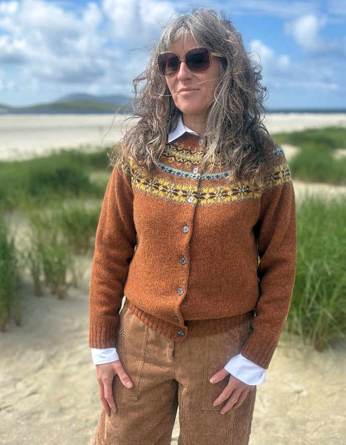 A woman wearing a rust-orange Fair Isle knit cardigan with a colorful patterned yoke, styled with brown corduroy pants and sunglasses, standing on a sandy beach dune with grass and a blue sky in the background.