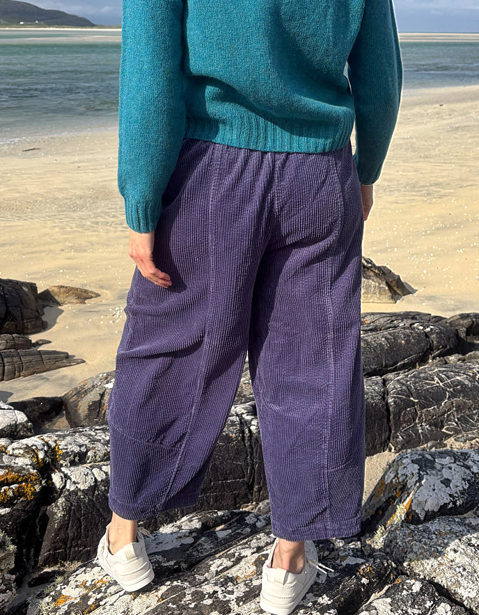 A person standing on a rocky beach wearing loose, wide-leg purple corduroy pants, a teal sweater, and white sneakers, with sand and water in the background.