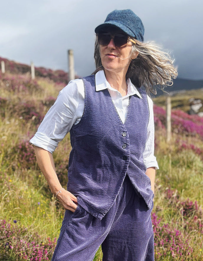A woman wearing a cord purple vest and matching trousers over a white collared shirt, standing outdoors in a grassy field with purple flowers under a cloudy sky.