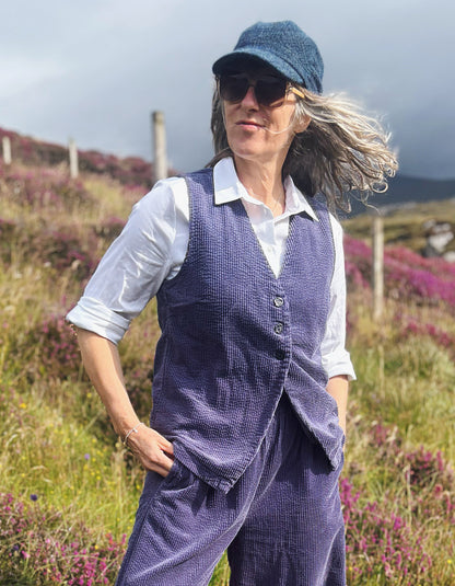 A woman wearing a cord purple vest and matching trousers over a white collared shirt, standing outdoors in a grassy field with purple flowers under a cloudy sky.