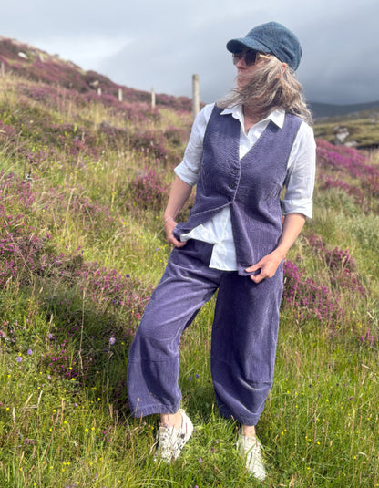 A woman stands in a grassy, heather-covered outdoor landscape wearing a matching slate blue corduroy vest and wide-leg trousers set, a white button-up shirt, a blue cap, and white sneakers.