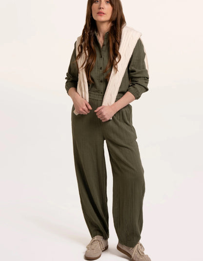 A woman modelling an olive green checked barrel leg trousers with a cream ribbed knit vest layered over it, paired with beige sneakers, against a plain white studio background.