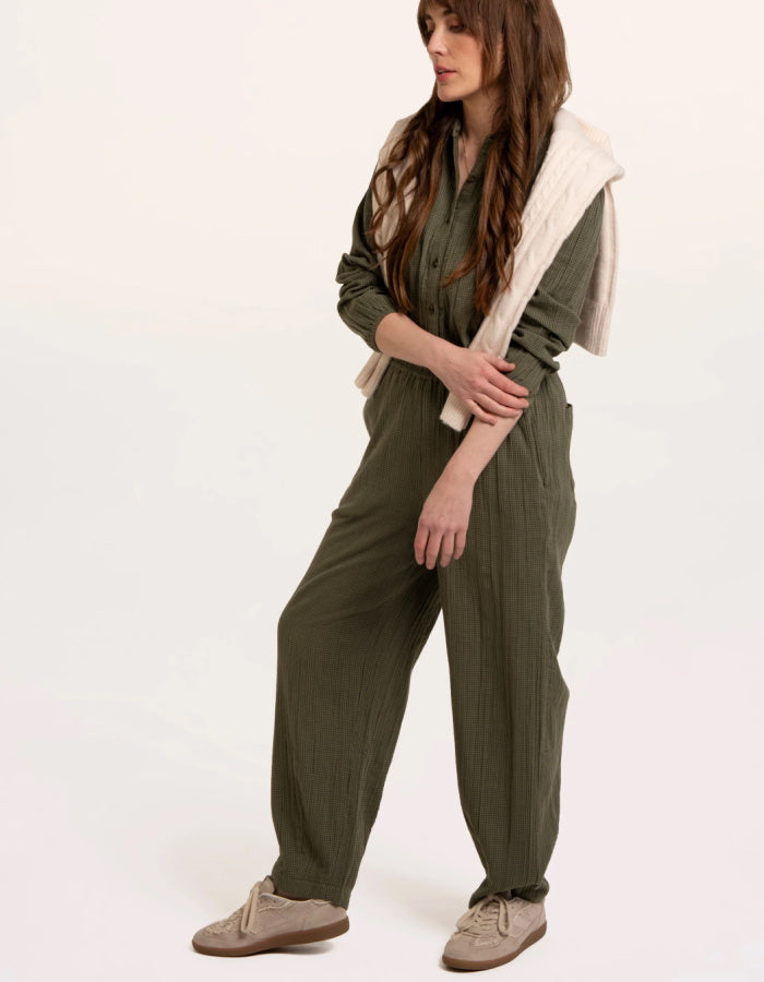 A woman modelling an olive green checked barrel leg trousers with a cream ribbed knit vest layered over it, paired with beige sneakers, against a plain white studio background.