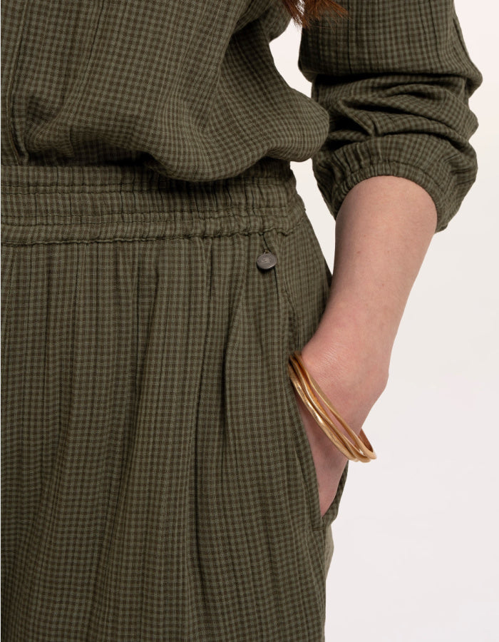 A woman modelling an olive green checked barrel leg trousers paired with beige sneakers, against a plain white studio background.