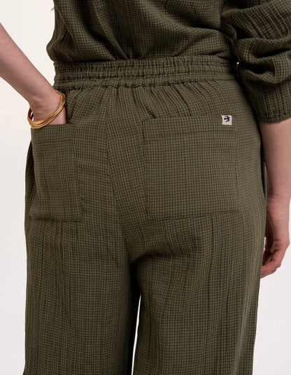 A woman modelling an olive green checked barrel leg trousers paired with beige sneakers, against a plain white studio background.