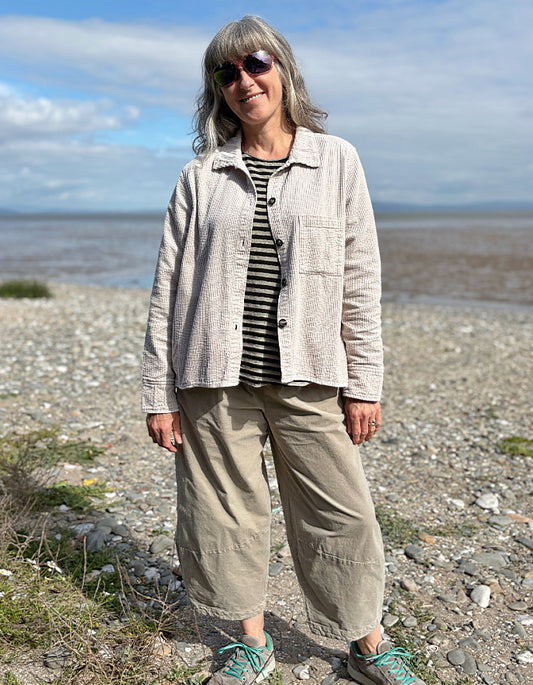 Woman standing on a pebbly beach wearing sunglasses and cream cord shirt, stripes t shirt and beige cord lantern trousers