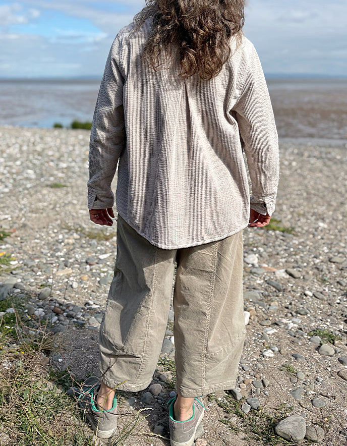 Woman standing on a pebbly beach wearing sunglasses and cream cord shirt, stripes t shirt and beige cord lantern trousers