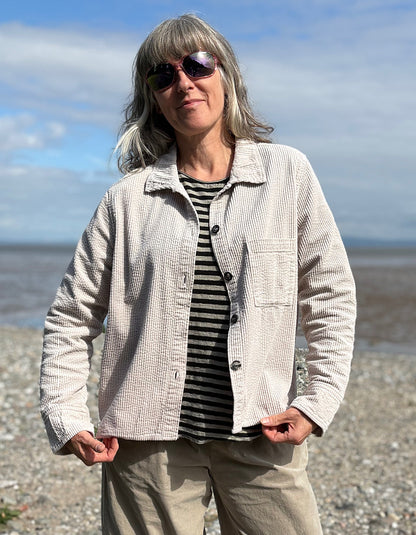 Woman standing on a pebbly beach wearing sunglasses and cream cord shirt, stripes t shirt and beige cord lantern trousers