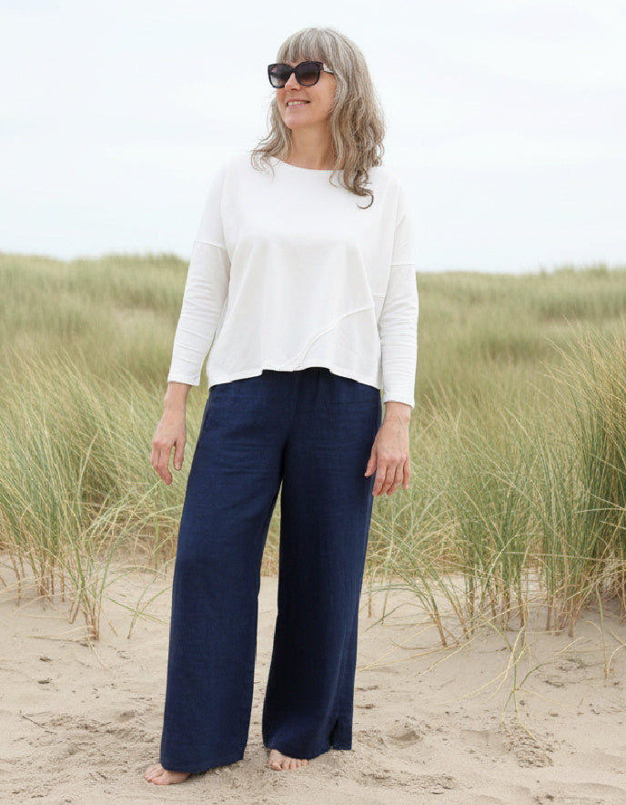 Woman wearing a white top and sunglasses on a sandy beach with grasses.