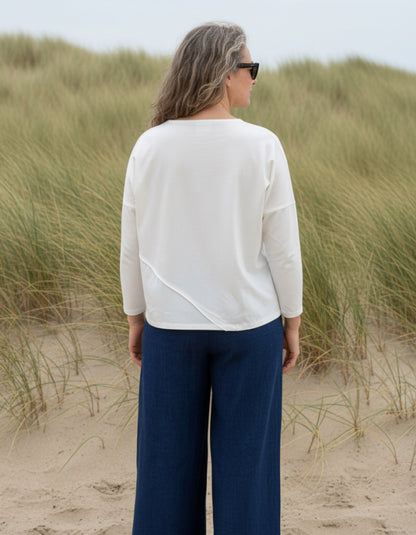 Person standing on a sandy beach with grassy dunes, wearing a white top and blue pants.