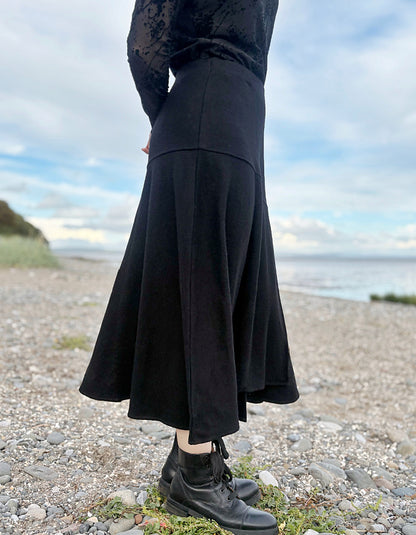 Person wearing a black lace top and black wool skirt standing on a pebbly beach with ocean and sky in the background.