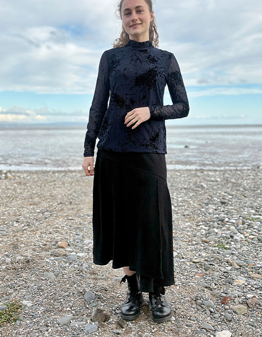 Person wearing a black lace top and black wool skirt standing on a pebbly beach with ocean and sky in the background.