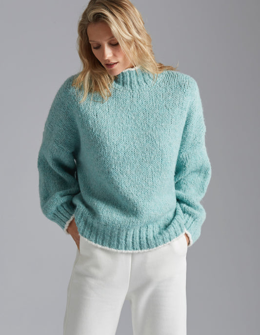 Woman wearing a teal sweater and white pants against a gray background