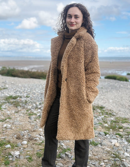 A woman with curly brown hair stands on a pebble beach wearing a long tan teddy coat over a brown turtleneck and dark plaid trousers, with a cloudy blue sky in the background.