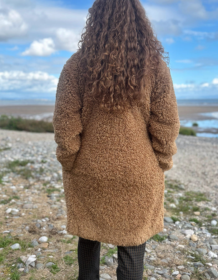 A person viewed from behind wearing a brown teddy fleece coat and black plaid trousers, standing on a rocky shoreline with water and a cloudy blue sky in the background.
