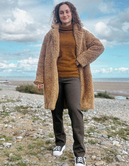 A woman stands outdoors on a rocky shoreline wearing a tan teddy-fleece coat over a rust-brown turtleneck sweater, dark patterned trousers, and white sneakers under a cloudy blue sky.