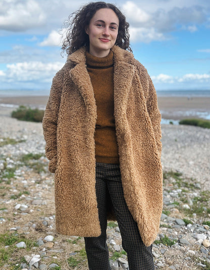 A woman with curly dark hair stands outdoors on a rocky shoreline wearing a long tan teddy-fleece coat over a brown turtleneck sweater and dark plaid trousers, with a cloudy blue sky in the background.