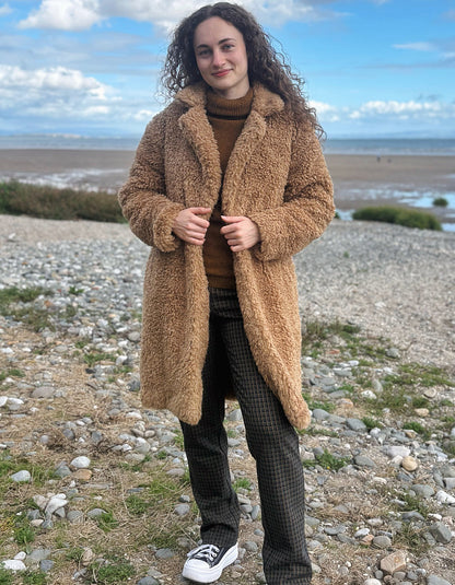 A woman wearing a long tan teddy coat over a brown turtleneck, dark plaid trousers, and white sneakers, standing on a rocky beach with the sea and blue sky in the background.