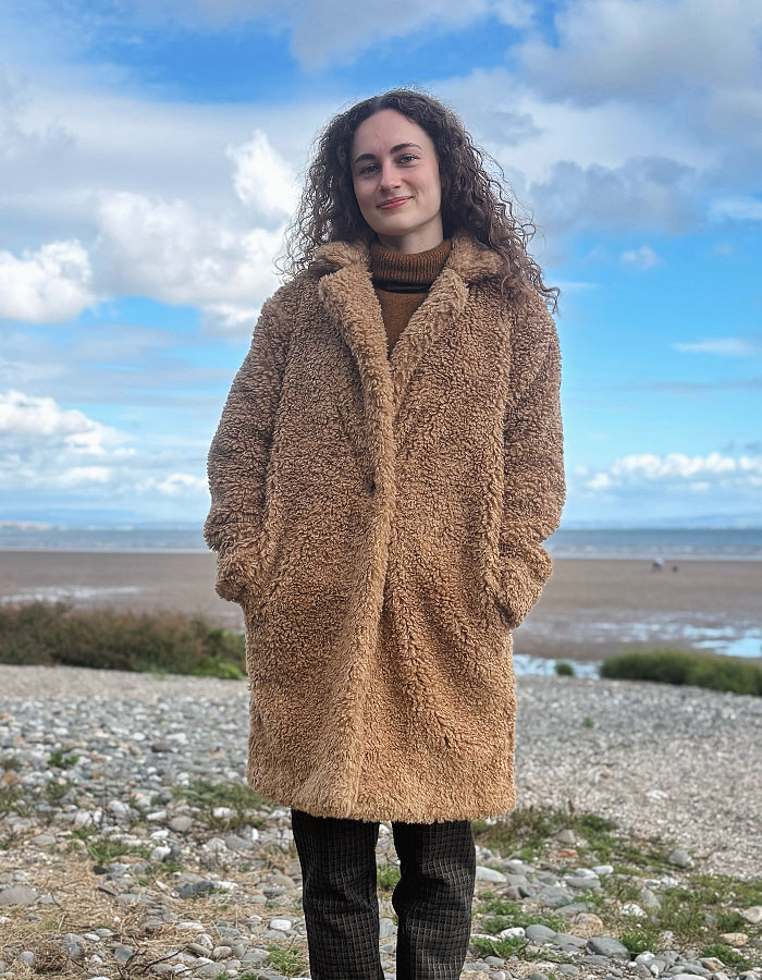 A woman with curly brown hair stands on a rocky beach wearing a long, plush camel-brown teddy coat over a brown turtleneck and dark plaid trousers, with a blue sky and clouds in the background.