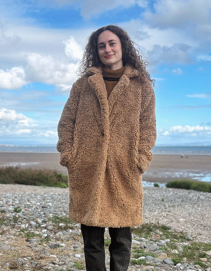 A woman with curly brown hair stands on a rocky beach wearing a long, plush camel-brown teddy coat over a brown turtleneck and dark plaid trousers, with a blue sky and clouds in the background.